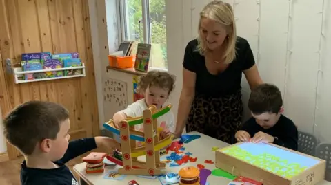 A woman with shoulder length blonde hair, a black short sleeved top and a leopard print skirt stands over a table where three young children are playing. One little boy is playing with some cars. A little girl is helping him. And another boy is playing with letters on a light box.
