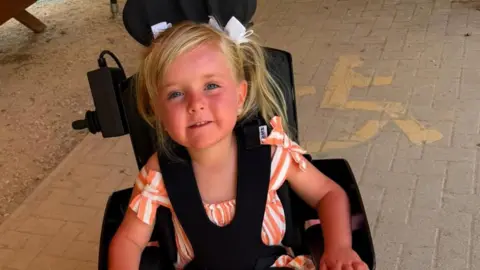 Warren Davies A young girl with blonde hair wearing a striped orange and white playsuit. She is sat in a black wheelchair with black straps holding her in. She is smiling and look at the camera.