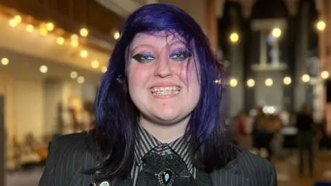 BBC Lexie has dark, shoulder‑length hair and stands in a large hall decorated with string lights. They are wearing a dark pinstripe jacket with multiple badges and a ruffled shirt. Rows of chairs and a tall arched structure are visible behind them.