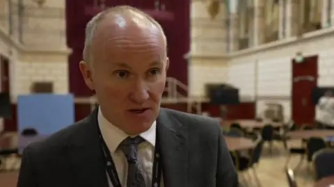 Det Supt Glenn with short fair hair wearing a grey jacket and blue tie with a blue lanyard. There are tables and chairs behind him as well as a stage.