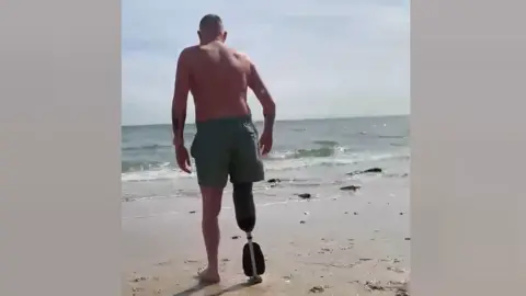 Alex Young walks away from the camera on a sandy beach towards the sea. He wears swimming shorts and a prosthetic right limb.
