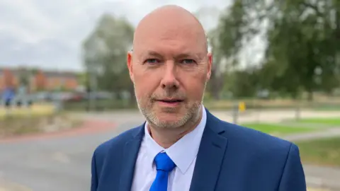 A head and shoulders image of a man wearing a dark blue suit jacket, white shirt and blue tie. He has a grey beard and blue eyes.