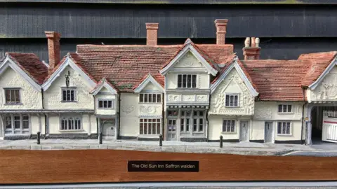 Simon Evans A model of an inn that has been placed on a wooden plinth. The building is cream and in a medieval style. The name of the pub and its location is written on a black plaque on the front of the wooden plinth. 
