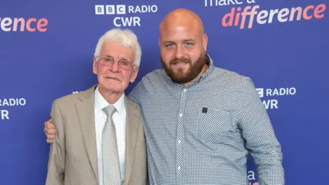 Mark Pemberton Steve is standing on the right wearing a blue and white shirt. He has a bald head. He has his arm around a man with white hair and glasses, who is wearing a suit