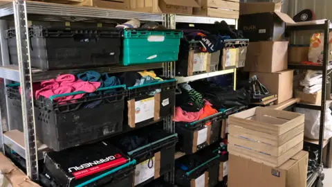 An indoor storage or stockroom space. There is a tall, industrial-style metal shelving unit with multiple shelves. Each shelf holds several large plastic crates and bins, most of them black or dark-coloured, with a few teal or green bins mixed in. These crates are densely packed with folded clothing.