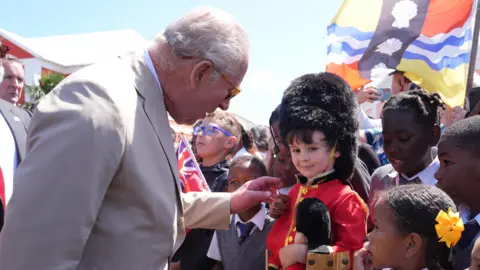 Reuters King Charles III speaks to a little boy in a guardsman outfit. The little boy is wearing a teddy bear that also wears the outfit.