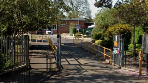 A path with railings on either side leading to a single-storey brick school building. There are trees and shrubs lining the front gates of the premises