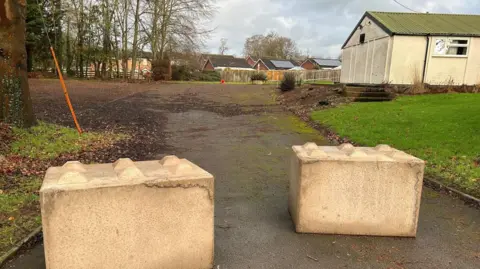 Two large concrete blocks at the entrance to a muddy car park with a white-walled building in the background
