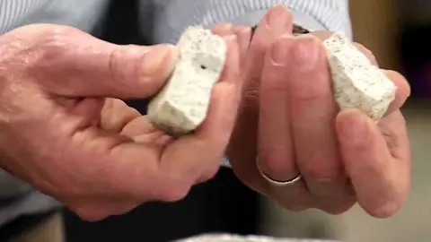 Close-up of a person holding two broken pieces of light grey, porous construction material above a work surface. The material's rough texture and internal structure are clearly visible, suggesting a demonstration or examination of a building product.