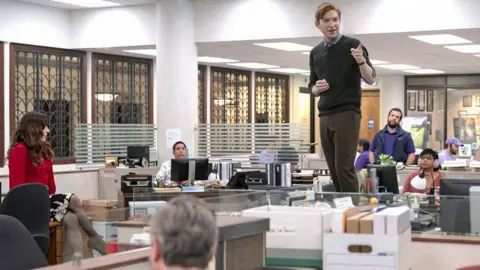 Peacock Domhnall Gleeson standing on a table in an office in character as Ned Sampson