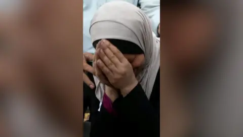 A woman in a headscarf clasps her hands in anguish.