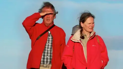 Miranda Mugford and her husband Mike standing next to each other. The are both wearing red outdoor-type coats. Mike has his right hand above his eyes, to avoid sun glare.