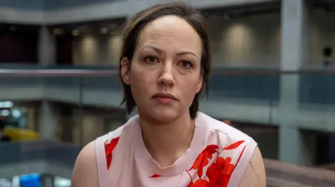 A woman with short, brown hair, wearing a pink and red floral sleeveless top, looks into the camera with a neutral expression