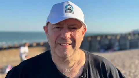 Karl is wearing a Trunks and Hunks hat. He is standing on the beach and smiling to the camera.