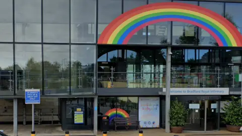 Google A glass fronted building with a revolving door on the right side. Above the door is a sign that reads Welcome to Bradford Royal Infirmary. A large rainbow decal stretches across part of the second floor. Outside the building there is a bench, two plants in pots and a sign that says 20 minute pick up and drop off only.