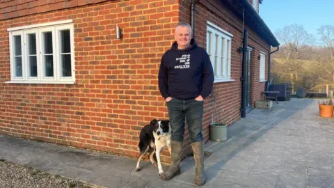 Simon Broad (a man in a black hoodie with dark jeans and wellington boots on)  with a black and white dog next to him stands in front of a house