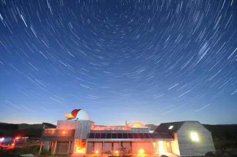 Marc Charron A brightly lit observatory building beneath a swirling starry sky