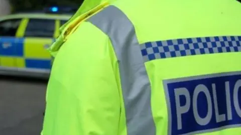 The image is the upper back and shoulder of a man wearing a bright fluorescent yellow police jacket. The jacket is neon yellow with reflective silver strips. There is a dark blue rectangular patch with the word POLICE in white capital letters. In the background, slightly out of focus, there is a marked police car.