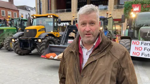 Richard Wheeler has grey hair and facial hair. He is wearing a brown jacket and is standing in front of parked tractors in a square. They have signs that read "no farming, no food, no future."