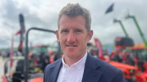 Man in suit jacket and open shirt standing in front of farm machinery