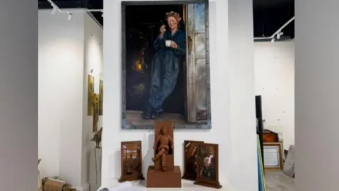 A large painting of a female steelworker leaning against a door frame is attached to a wall inside Sunderland's 24NE Gallery. The painting is in dark colours and shows the woman holding a cup in one hand a cigarette in another. The statue maquette is on a table beneath the painting. It is bronze-coloured and shows a female steelworker sitting with her legs crossed.