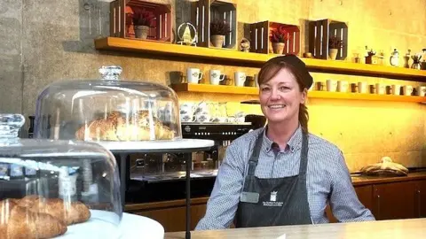 A woman stands behind a café counter with shelves of cups, jars, and decor arranged neatly against a warm-toned wall. Glass domes in the foreground display pastries, including croissants, adding to the cosy bakery atmosphere.