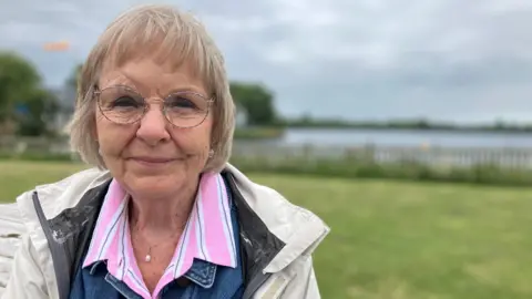 Jackie Heywood is an older women with fair, blonde, short hair. She is wearing round framed glasses, a pink shirt under a beige rain jacket. She is stood with a lake behind her. 