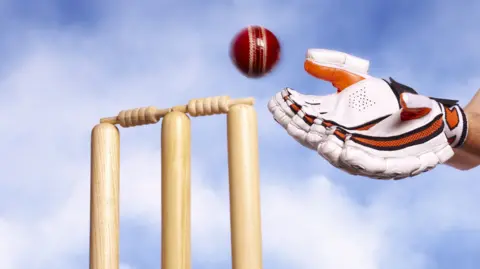 A gloved hand attempts to catch a cricket ball as it nears the stumps