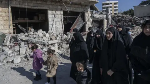 Two little girls in coats hold hands as they walk along a street filled with rubble with a group of women who are wearing hijabs 