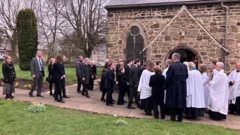 Mourners entering St Mary Magdalene Church in Trimdon. The small sandstone church is surrounded by green grass. Mourners dressed in black and a number of people wearing white clergy gowns are queueing to enter the church.