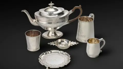 Silver items including a plate, jug, and three cups, against a dark background.