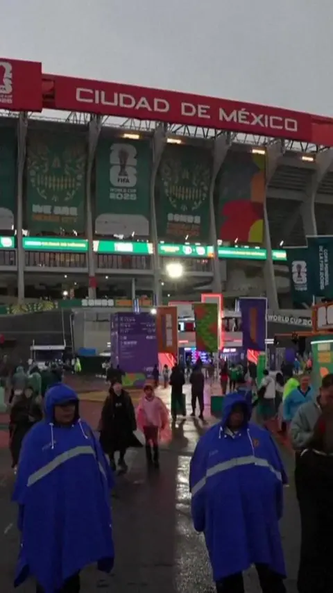 A general view outside Estadio Azteca. People are scattered across the site. The exterior of the stadium is lit up with screens.
