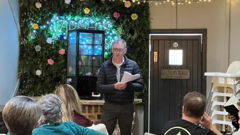 Mark Terry-Lush wears a black coat and trousers and a white shirt as he talks to people who are sitting facing him. Terry-Lush wears glasses and has short grey hair and is holding a stack of papers. He is standing in front of a leaf wall with roses in it and there is a door with laundry room written on it to his side. 