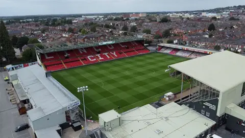 BBC Aerial view of Sincil Bank at home in Lincoln City. It has four pillars and the city of Lincoln floods in the background. The main stand has white seats that train Lincoln City on red seats around it.