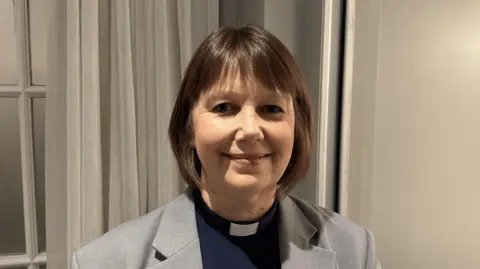 A woman with short brown hair wearing a grey blazer and a dark blue shirt with a vicars white dog collar. 