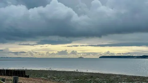 Janeybeth A beach scene but the sky is dark grey with ominous looking clouds. There is a line of lighter sky in the distance. A ferry can been seen on the water in the far distance