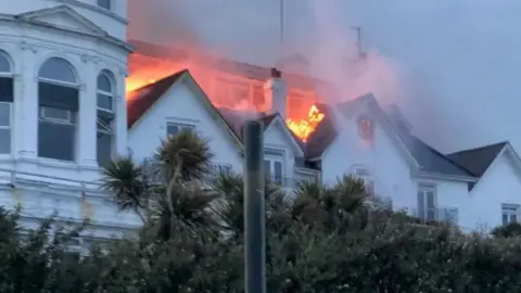 Sandown Hub Large white building with pitched roofs and orange glow from flames in its centre 