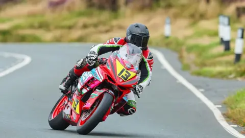 Manx Grand Prix Louis O’Regan riding a motorbike on the Mountain Course. His bike is red with the number 13 in black writing on a yellow plate and he is wearing red, white, green and black leathers and a black helmet.