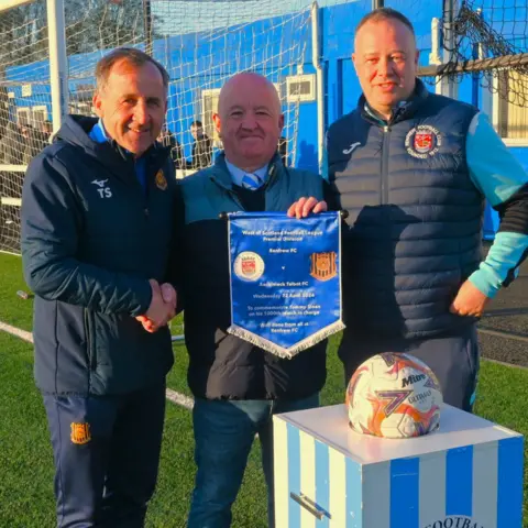 Deltamike Images Three men stand together at a football ground in front of a goal net and blue building; two shake hands while the middle man holds a commemorative pennant marking a West of Scotland Football League Premier Division match between Renfrew FC and Auchinleck Talbot FC.