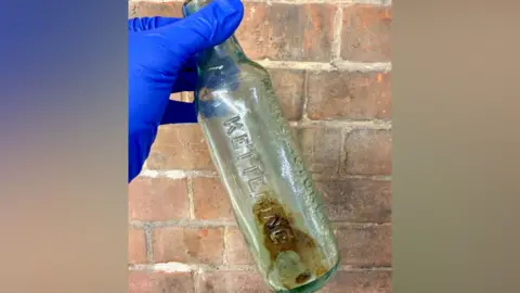 North Northamptonshire Council An old empty glass bottle, being held up by a person wearing blue gloves. The bottle is being held up in front of a brick wall. 