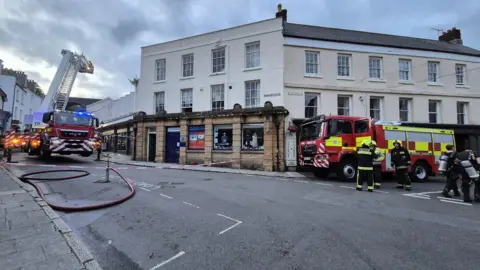 On Market Street there are two fire engines visible. One is resting on its hydraulic supports and has a ladder extending from it. the other has firefighters wearing breathing apparatus standing near it. There is incident scene tape stretched across the street and a large and long hosepipe in the foreground.