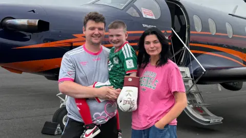 Máirtín McGabhann Dáithí is pictured with his mum and dad. He's dressed in a green sports jersey with a boxing glove on one hand and is in his dad's arms. Behind them is a plane.