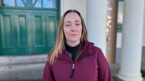 A woman stands outside a Victorian style building. There are columns and green doors in the background. You can see part of a sign that reads visitor centre. She is wearing a maroon coloured winter coat. 