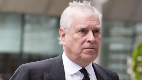 Andrew Mountbatten-Windsor is an older man with white, thinning hair. He is outdoors in a city street and wearing a grey suit jacket over a white shirt with a black tie.