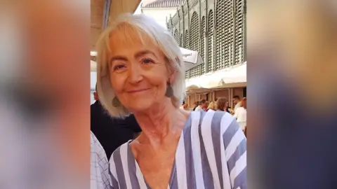 A woman smiles into the camera while sitting outside at a café. She is wearing a blue and white stripped top and has bobbed blond hair and is wearing earrings. The street is busy with people having something to eat.  