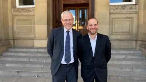 The pair are stood on the steps of the council house. Both are wearing dark suits. Harmer has a tie on, Pritchard does not. They are both smiling at the camera.