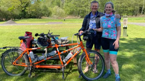 Chris and Nicky Clark Chris and Nicky Clark standing beside their tandem, Chris has his arm around Nicky and they are both wearing brightly coloured cycling shirts and black shorts. The bike is propped up beside a picnic table in a field with woodland behind. It is a sunny day. All their luggage is stacked on the table