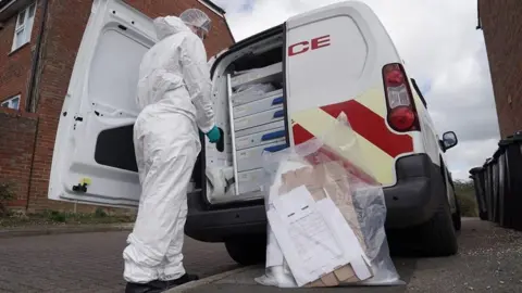 Essex Police A forensics officer in a white forensics suit with black shoes, stood at the back of a white police van. One of the doors of the van is open, with grey shelving inside. A transparent plastic bag is on the floor next to the forensics officer, containing several items.