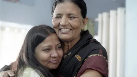 Anila - dressed in a black and maroon sari - and Monica hugging and smiling 