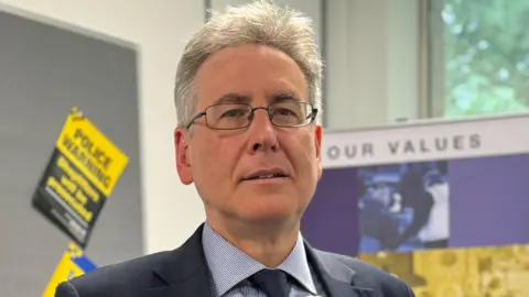 A man in a blue striped shirt, navy suit jacket and tie. He has white hair and is wearing glasses. He stands in a room with police leaflets pinned on boards behind him.